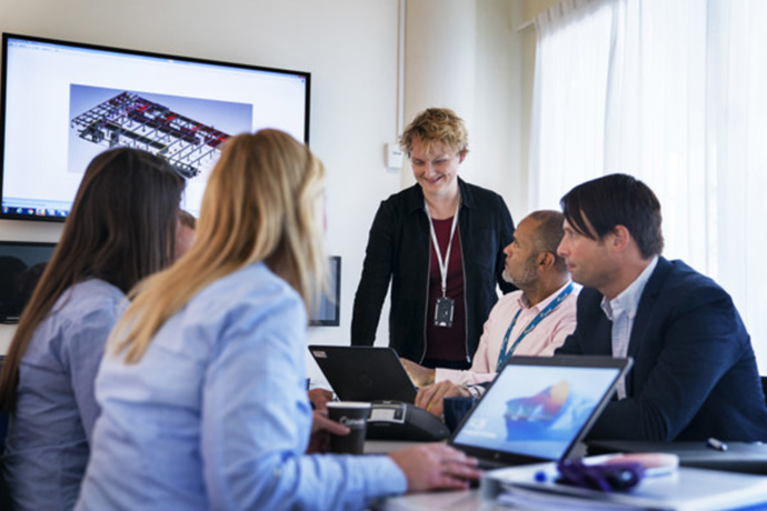 Several people are sitting around a table in a meeting room, with laptops and technical drawings on the screen during a project review.