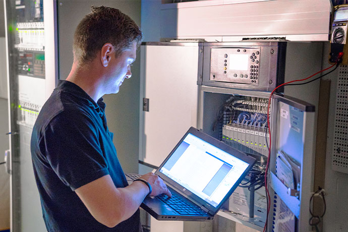 Person works with laptop connected to electrical and control systems in technical space, with open electrical cabinet and wiring.
