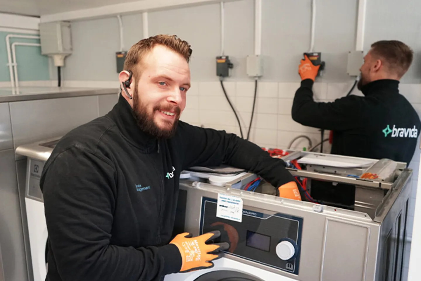 Bilden visar två servicetekniker som arbetar med att installera eller reparera tvättmaskiner. De bär svarta arbetskläder och orangea skyddshandskar.