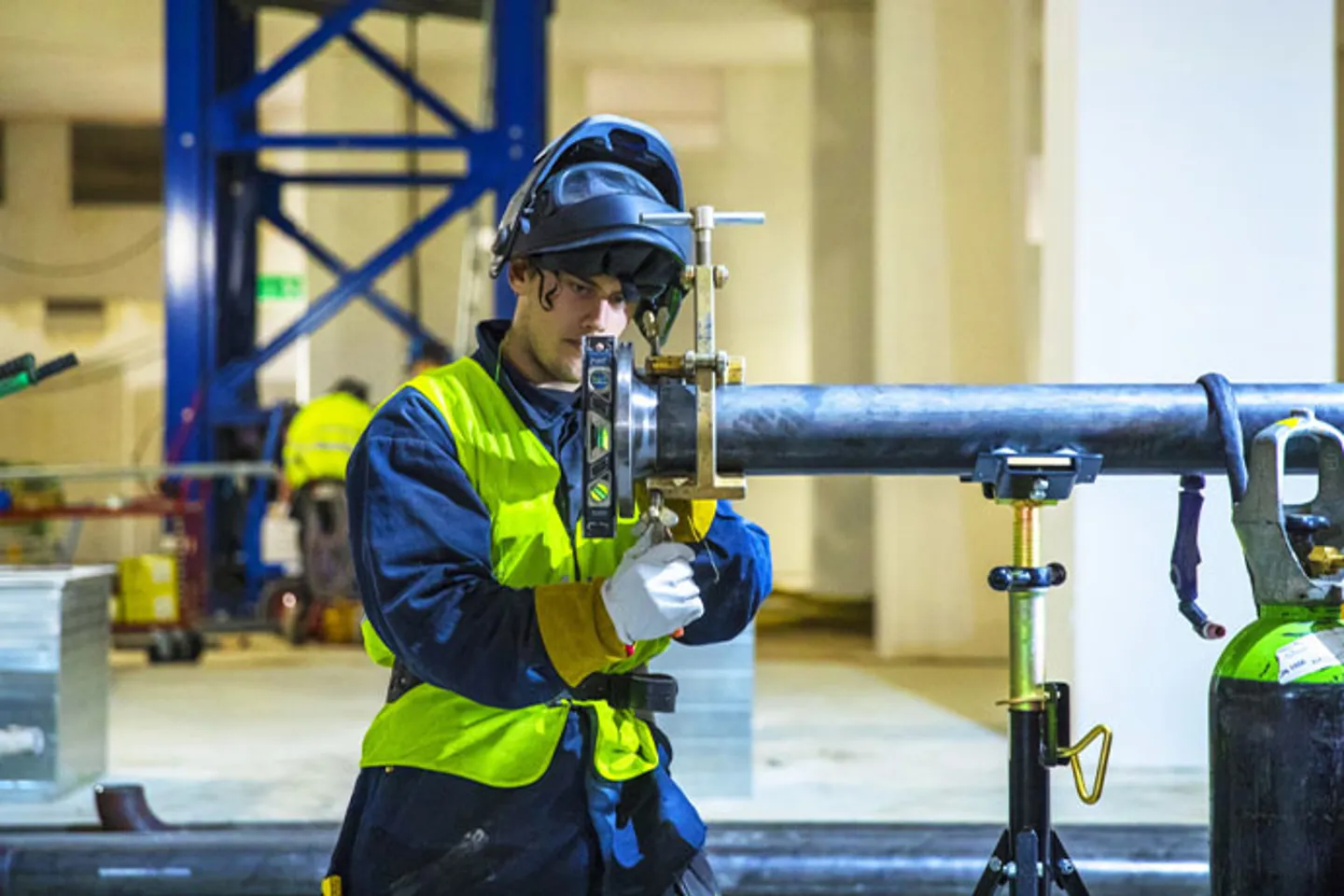 Bilden visar en VS-montör på en industriell arbetsplats. Personen bär skyddsutrustning, inklusive hjälm och varselväst, och är i färd med att arbeta med ett större rör som är uppställt på stöd.