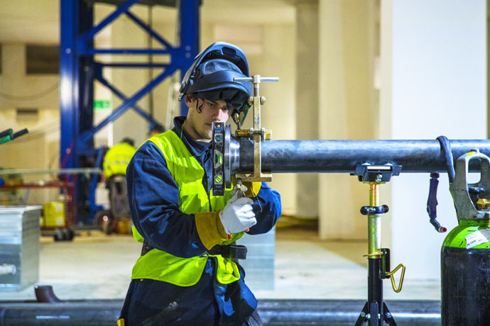 Bilden visar en VS-tekniker som arbetar med ett metallrör som är fastsatt med klämmor och ställningar i en industriell miljö. Personen bär: skyddshjälm, skyddsglasögon, handskar, varselväst.