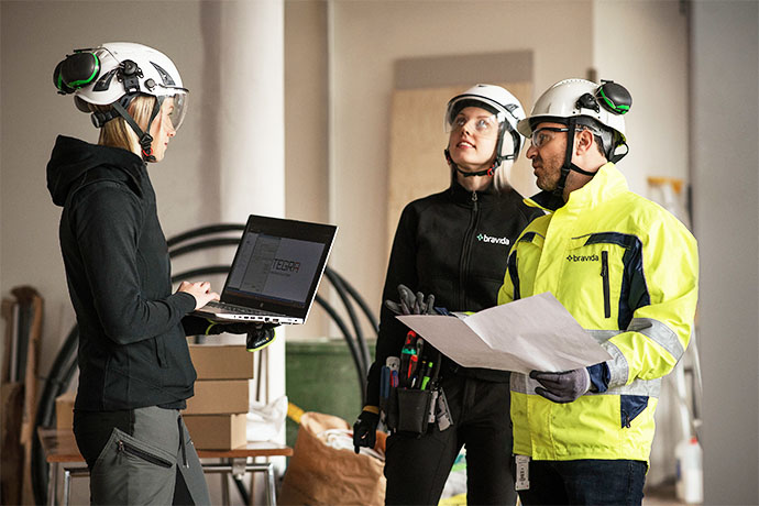 Three technicians in helmets stand in a construction environment and review drawings, while one uses a laptop for design and planning.