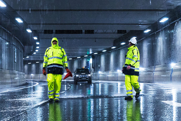 Bilden visar två personer i varselkläder som står i en upplyst, våt tunnel med ett fordon i bakgrunden.
