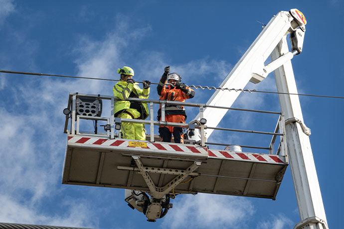 Bilden visar två personer i säkerhetsutrustning som står på en upphöjd plattform kopplad till en stor vit lyftkran. De arbetar med eller inspekterar luftledningar under en blå himmel, vilket tyder på underhålls- eller reparationsarbete.